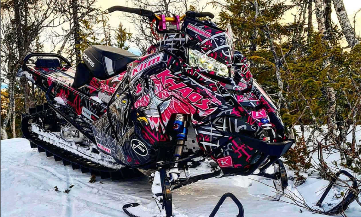 A snowmobile is parked in the woods, covered with a black and pink sled wrap. 