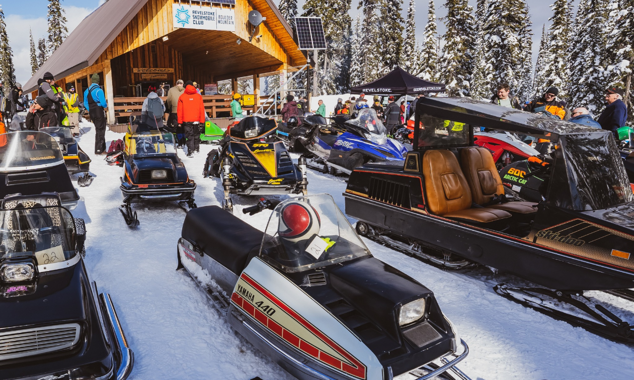 A collection of vintage snowmobiles is in front of a warm-up cabin. 