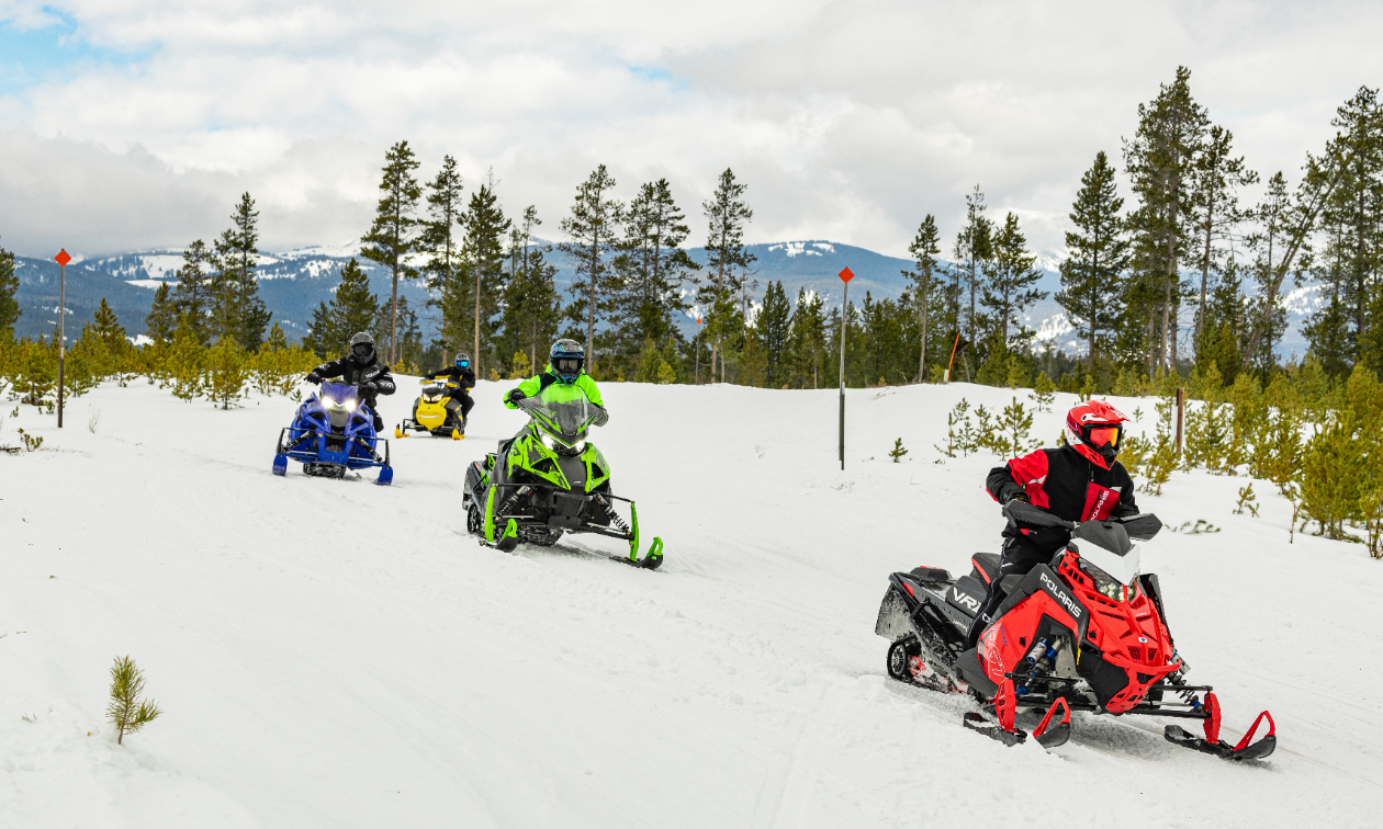 Four snowmobilers cruise around a corner. The sledder in front is all red. The next is green. Third place is blue. Yellow rounds out the rear. 