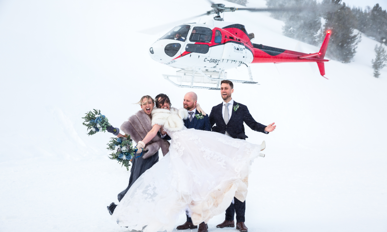 A helicopter flies behind a wedding-gown wearing Sarah Liebeknecht, formal-suited Damian Liebeknecht, and two more guests in the Purcell Mountains. 