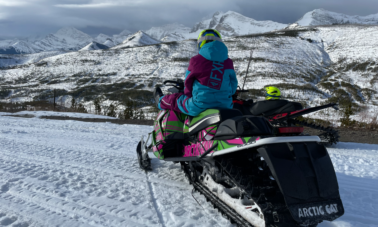 Madison Schwengler sits on her black and pink 2017 Arctic Cat M 6000 Mountain Cat 153 while looking at a snowy mountain horizon. 