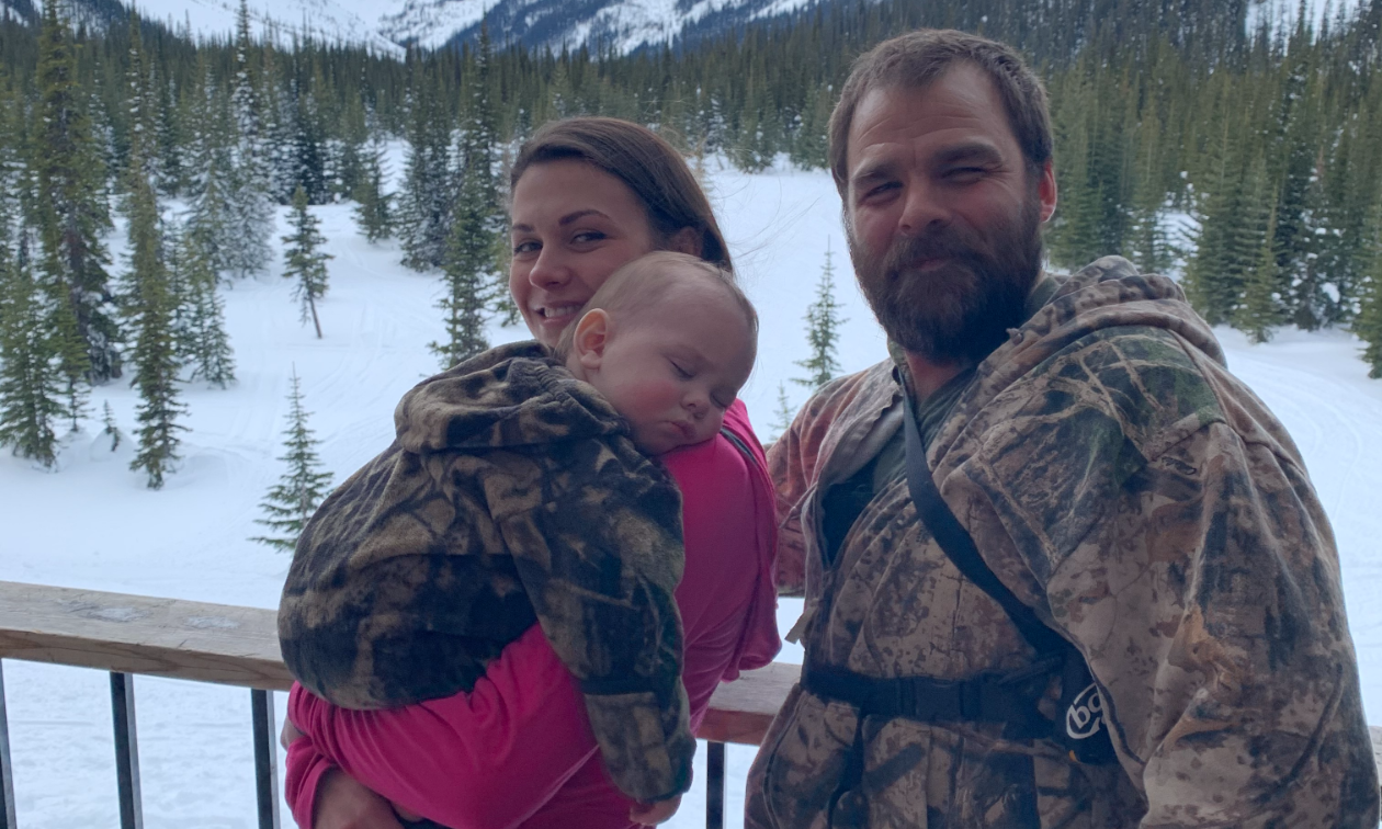 Bobby Chomica and Shelby Ingram hold their son, Mason Chomica, on a balcony of a warm-up shelter with snowy mountains in the distance.