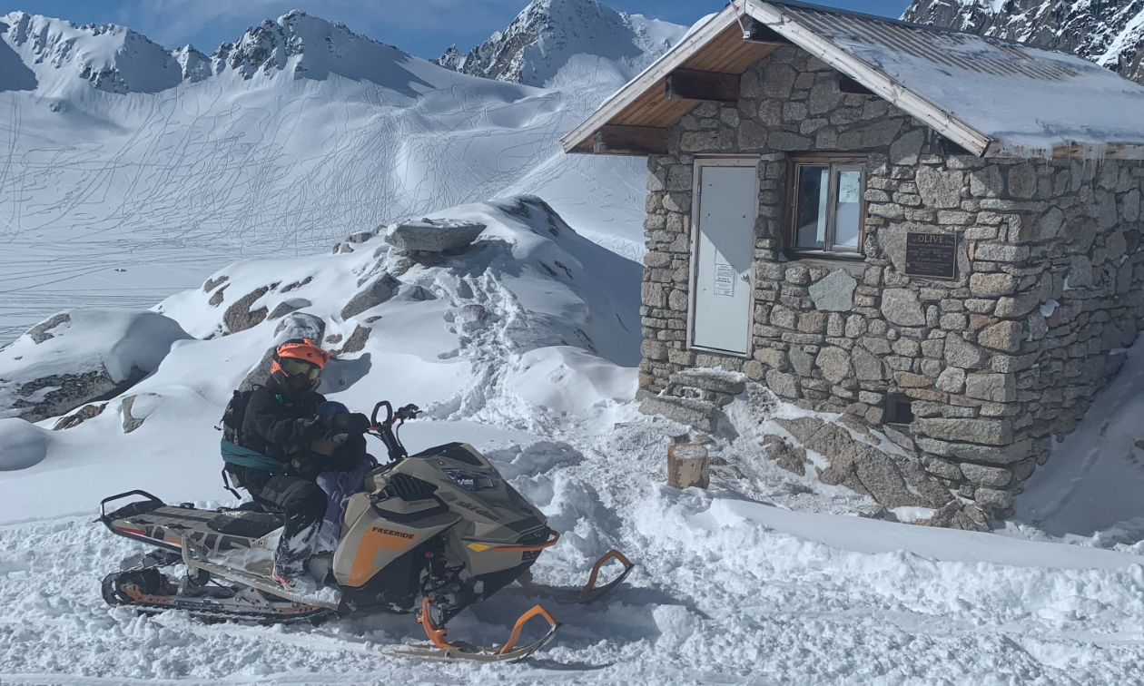 A snowmobile with two riders is parked in front of a stone warm-up shelter. 