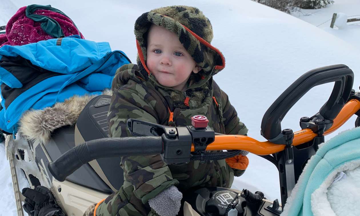 One-year-old Mason Chomica sits on a snowmobile wearing a thick green jacket. 