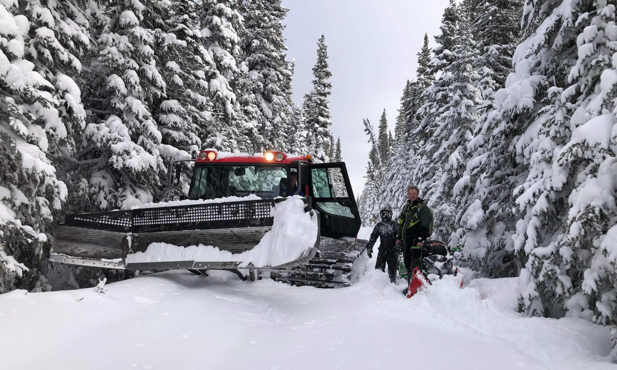 A snowmobile trail groomer is parked next to two men on a snowmobile trail. 