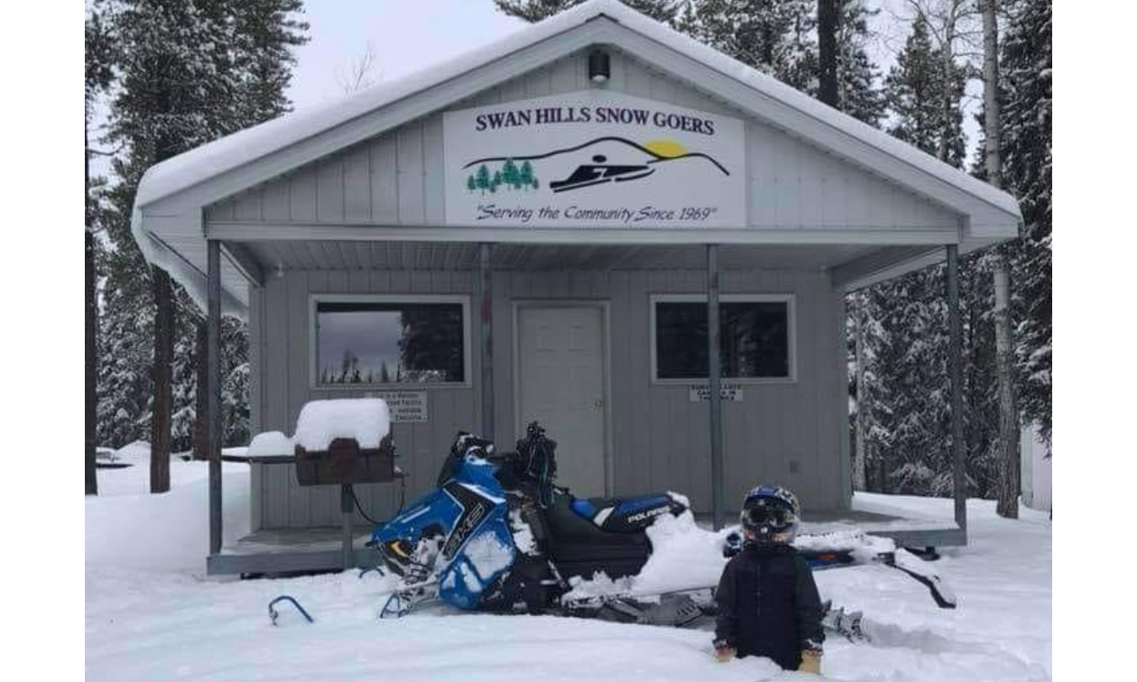 A boy sits in the snow in front of a snowmobile in front of a snowy warm-up shelter. 