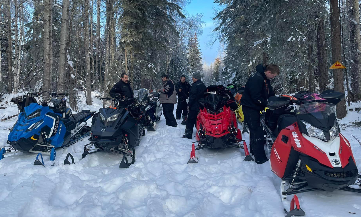 A group of snowmobilers takes a break on the trail between a row of trees on each side of the trail.
