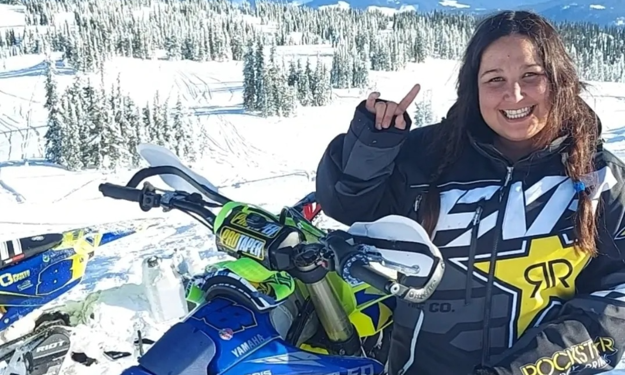 Tess Mason holds throws up the bull horns with her fingers while standing on a snowy mountain next to a Timbersled snow bike. 