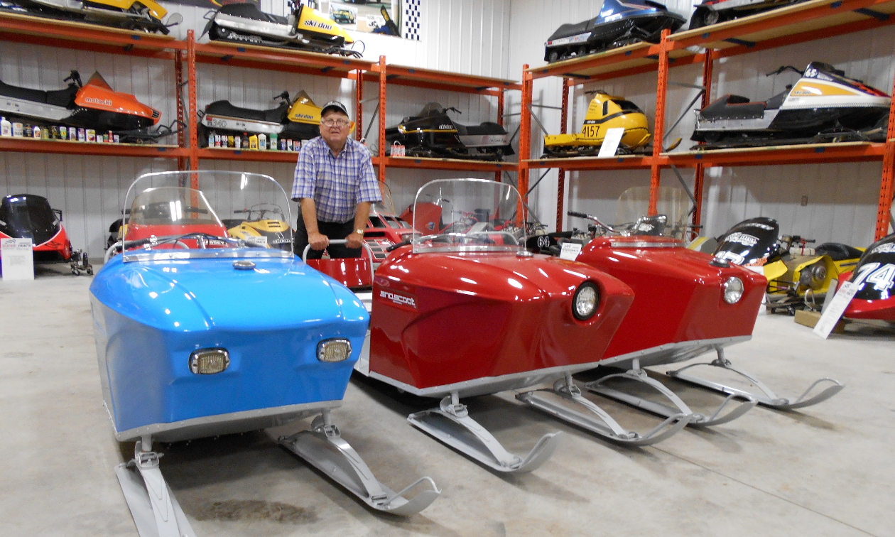 Art Bilous stands behind several Sno Scoot snowmobiles in a snowmobile museum.