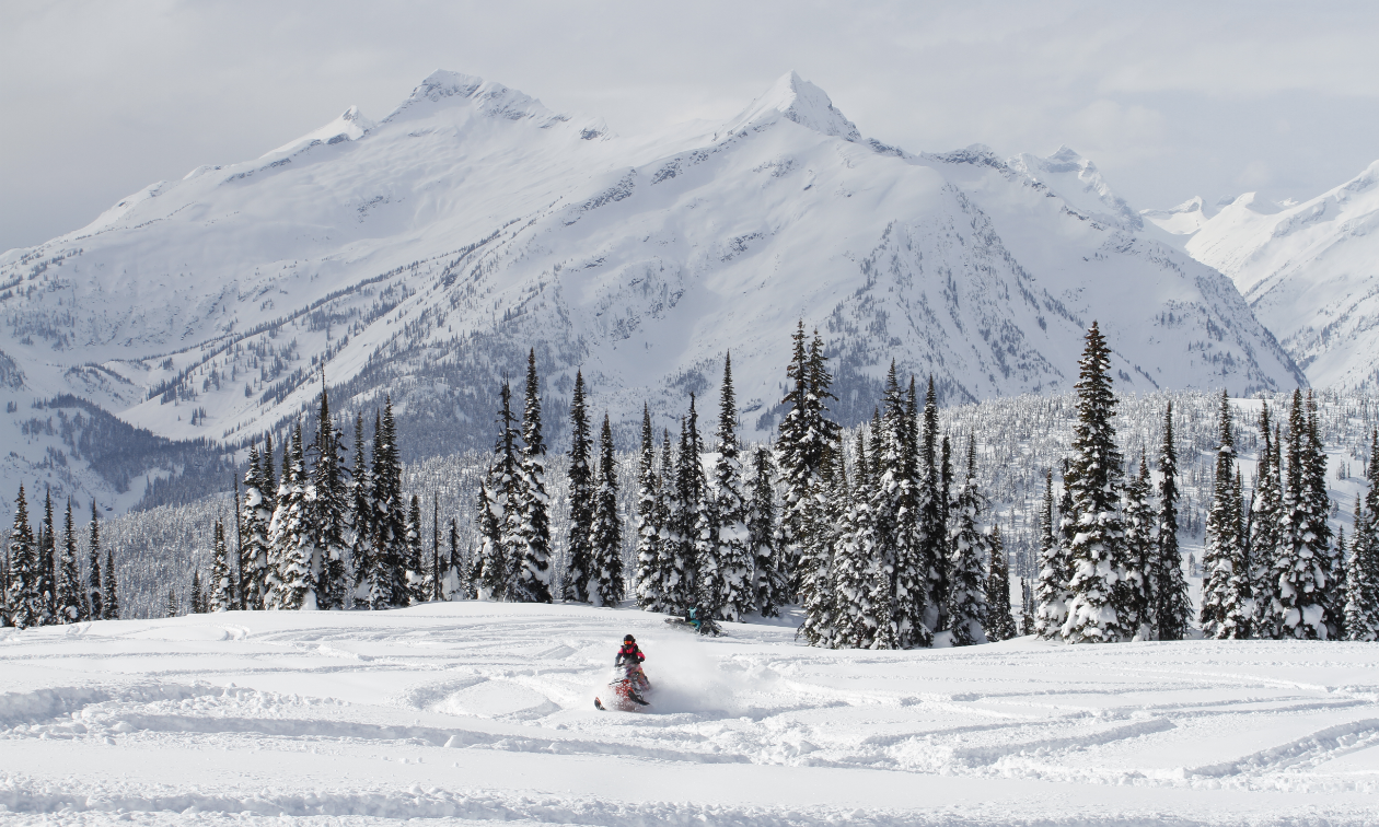 A snowmobiler carves through fresh snow as looming mountains tower in the distance. 