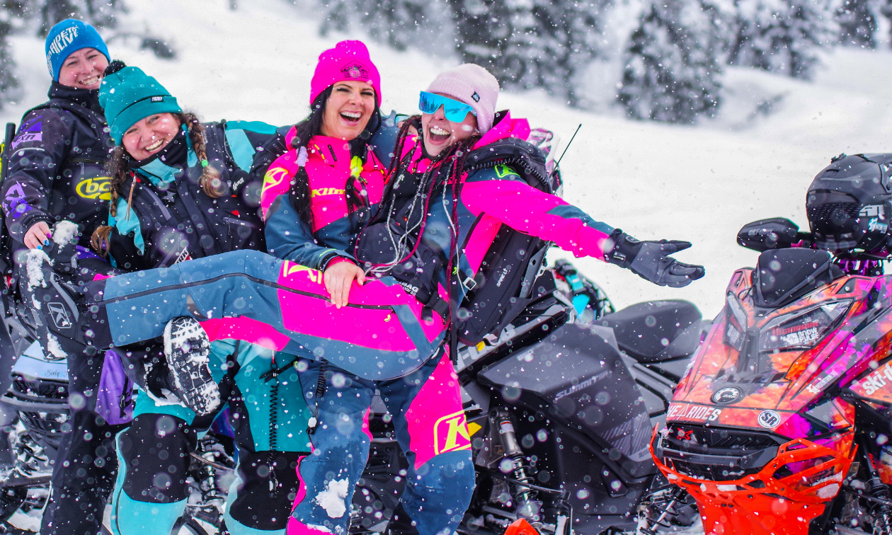 Taylor Craig-Gidney smiles as she is held by a fellow female snowmobiler and some other women laugh in the background