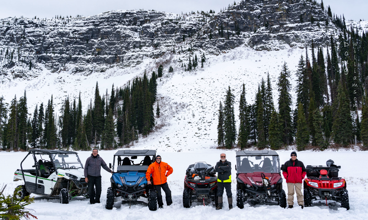 Five ATVers pose next to their ATVs at the base of a snowy mountain. 
