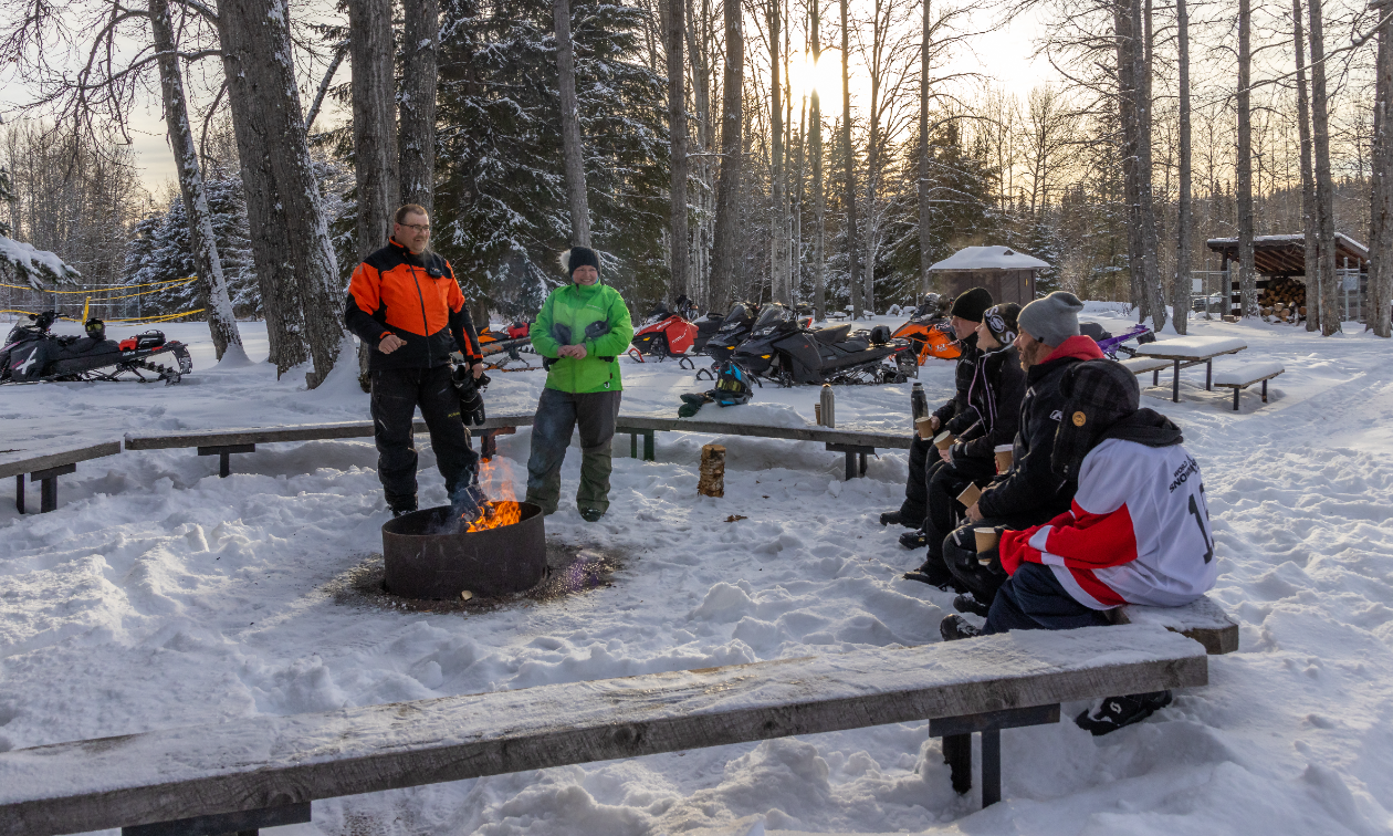 A group of snowmobilers sit on benches around a fire in wintertime. 