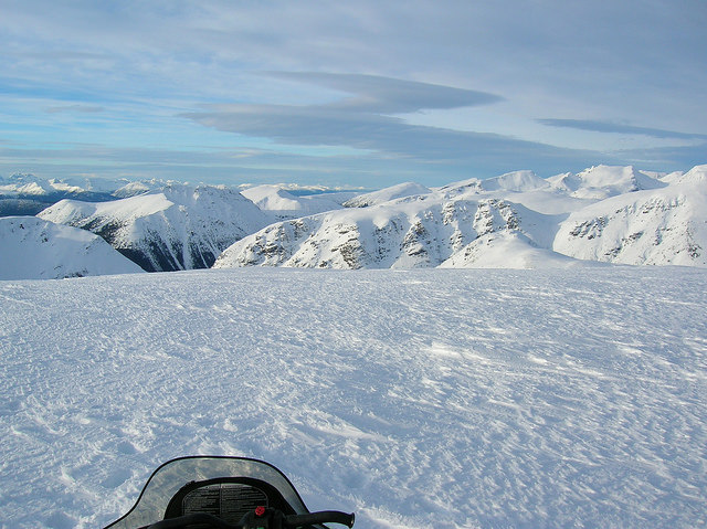 The windshield of a snowmobile is show in the forground and a stunning view of the Telkwa Range is off in the distance. 