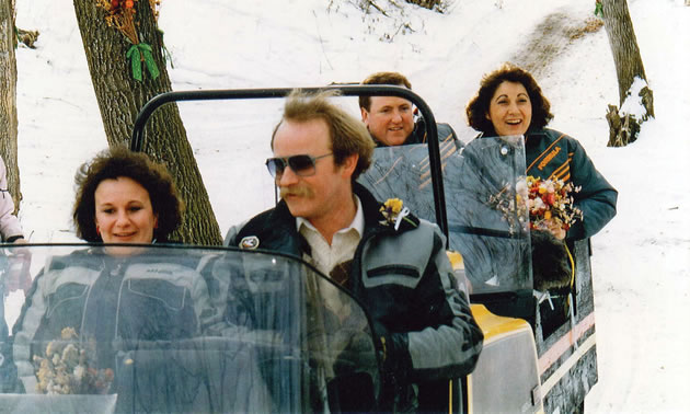 Photo of two people driving a two seater snowmobile and towing a couple behind in a cutter on their way to a wedding.