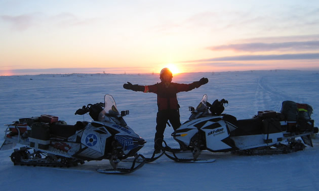 A guy standing between two white sleds with his arms open and the sun is setting behind him.