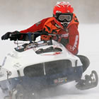 A close up of a man in a red suit driving a snowmobile around a racetrack. 