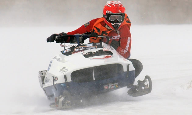 A close up of a man in a red suit driving a snowmobile around a racetrack. 