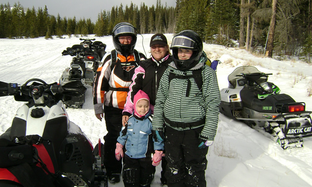 A grandmother, mother and her two daughters standing together dressed in snowmobile gear and they are surrounded by snowmobiles. 