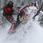 A woman with long black hair wearing a red outfit taking a jump on a red snowmobile.