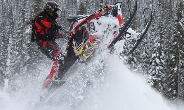 A woman with long black hair wearing a red outfit taking a jump on a red snowmobile.