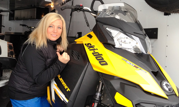 Photo of a blonde haired woman crouching beside a yellow and black snowmobile in an enclosed snowmobile trailer.