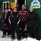 A group of riders in snowmobile gear stand outside a green corrugated cabin in the snow. 