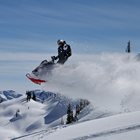 A sledder is air born, leaving a stream of powder behind against a blue sky and an array of mountains.