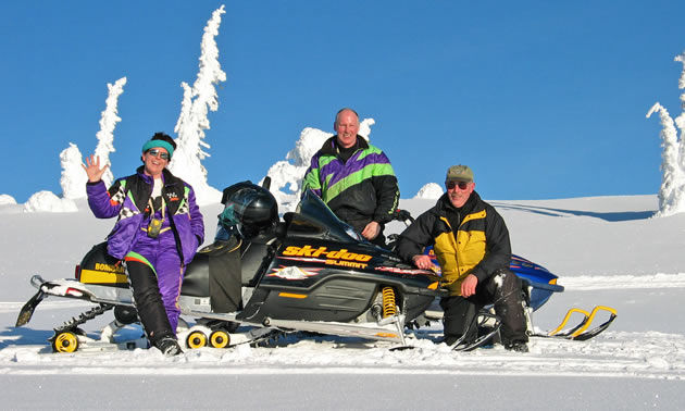 Three people on older snowmobiles. 