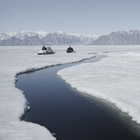 Photo of two sledders crossing ice in the Arctic near an open section of water.
