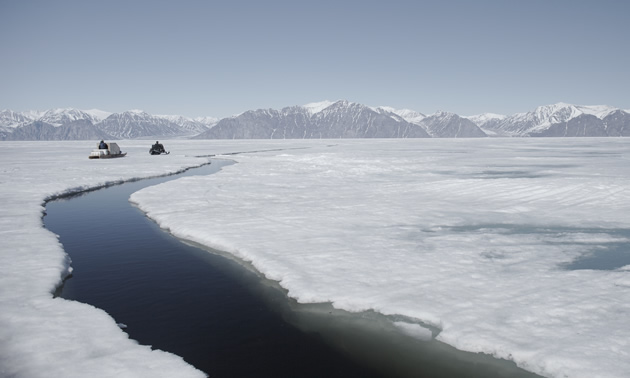 Photo of two sledders crossing ice in the Arctic near an open section of water.