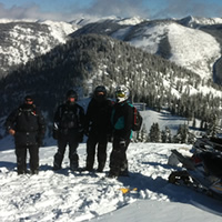 Four snowmobilers standing on top of a mountain by their snowmobiles. 
