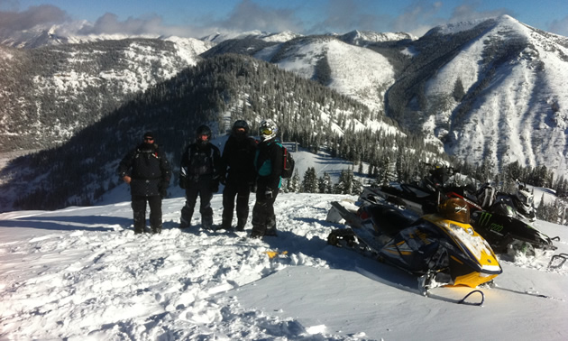 Four snowmobilers standing on top of a mountain by their snowmobiles. 