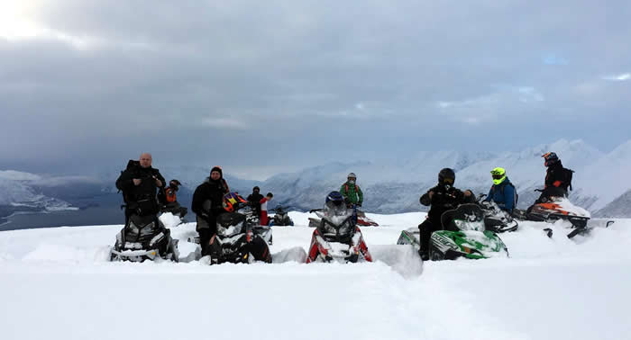 A group of snowmobilers stopped at the top of a mountain while breaking trail. 