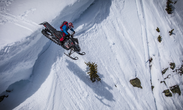 A man in a blue jacket and red pants dropping a black sled over a cornice. 