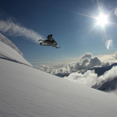A snowmobiler gets massive air off a mountain as the sun shines overhead. 