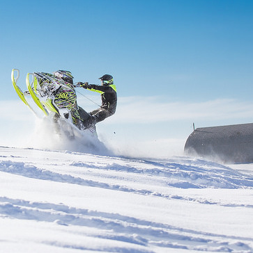 A snowmobiler does a wheelie. A gray barn is in the background.