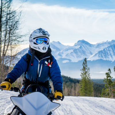 A snowmobiler looks on in the foreground with a horizon of mountain peaks in the background.