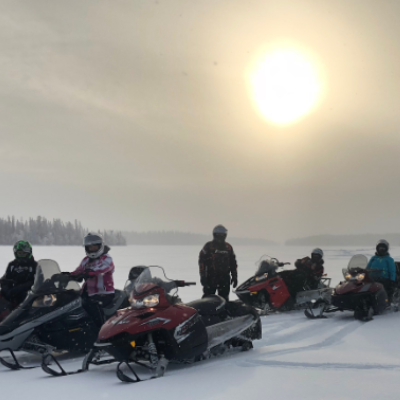Six snowmobilers stop on a flat snowy field near Flin Flon, Manitoba, with the sun shining overhead.