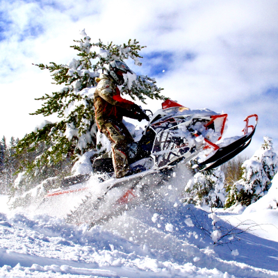 A snowmobiler gets air off a jump near a tree covered in snow.