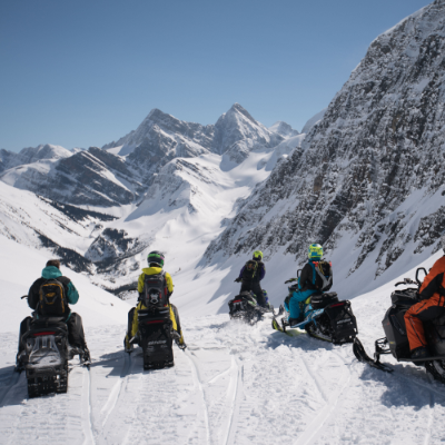 Six snowmobilers drive into a snowy valley.