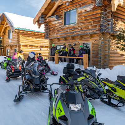 Snowmobiles are parked in front of a log cabin. People gather on the porch to visit with each other. 