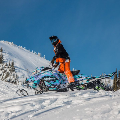 A man in orange pants stands up at the controls of a turquoise and purple snowmobile. 