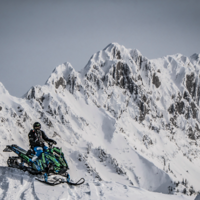 A man sits on his snowmobile amidst towering mountain peaks filled with snow. 