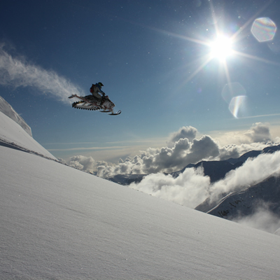 A snowmobiler gets massive air off a mountain as the sun shines overhead. 