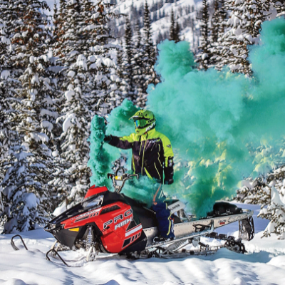 A woman stands on a snowmobile with a blue plume of smoke trailing behind her. 