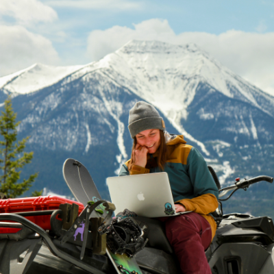 Brittney Dickson sits on her snowmobile while looking at her laptop in the mountains.