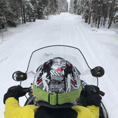 A snowmobiler looks ahead to a long stretch of groomed trail in between a line of trees.