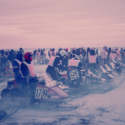 Snowmobile riders line up at a starting line in blistering cold conditions