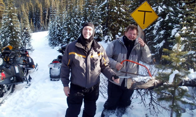 Two snowmobilers clearing a tree off the trail. 